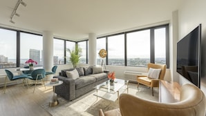 A spacious living room with a gray sofa, blue chairs, a coffee table, and a large TV. - Deedee's Sky Rise Apartments - Newport 2 (Jersey City)