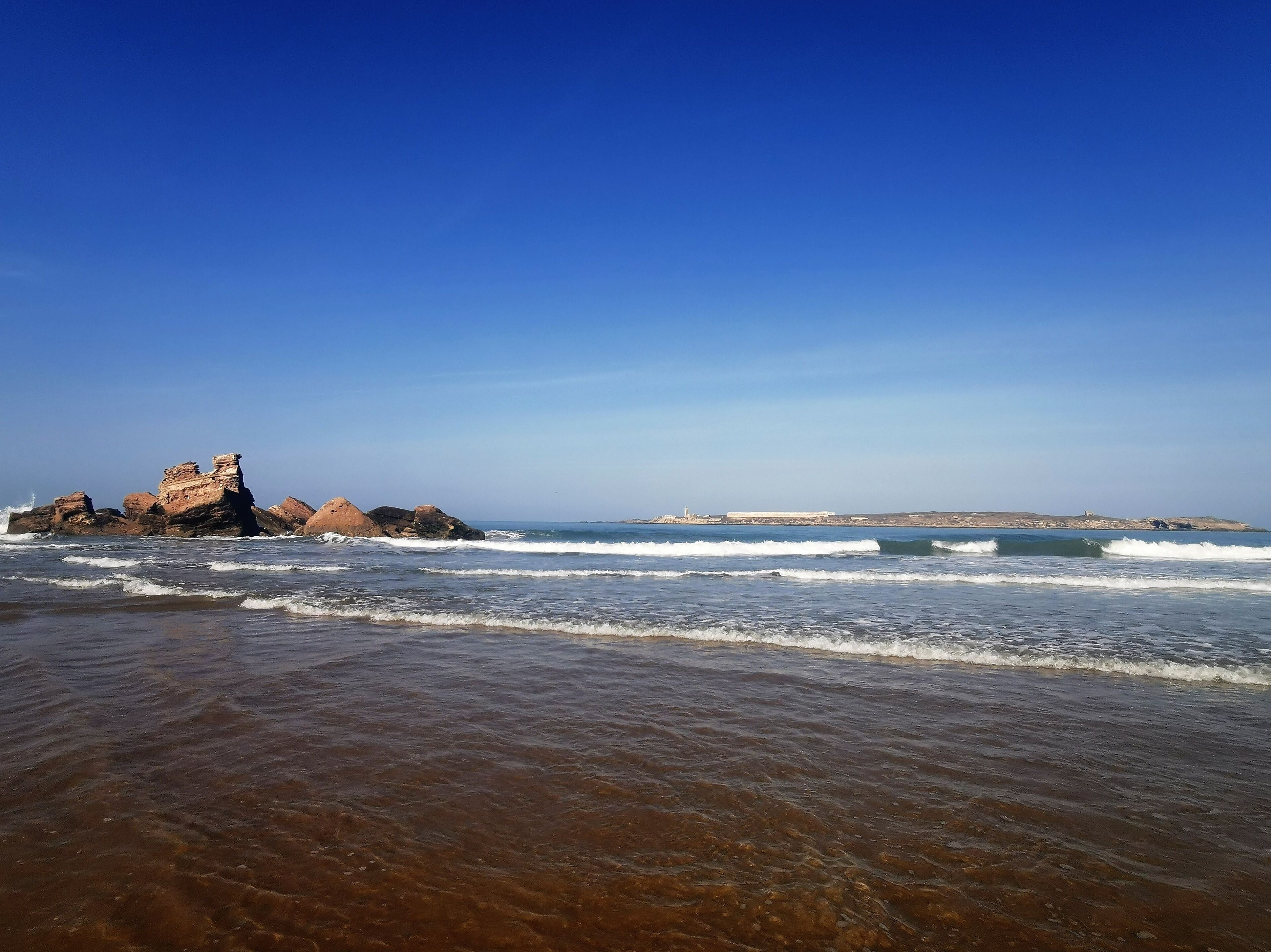 Une plage avec un ciel dégagé, des vagues et des formations rocheuses.