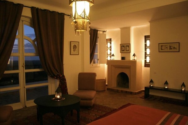 A cozy living room with a fireplace, a comfortable armchair, a small table with a candle, and a view through a large window with curtains. - Beautiful villa with sea view, large pool, Close to golf and the beach (Essaouira)