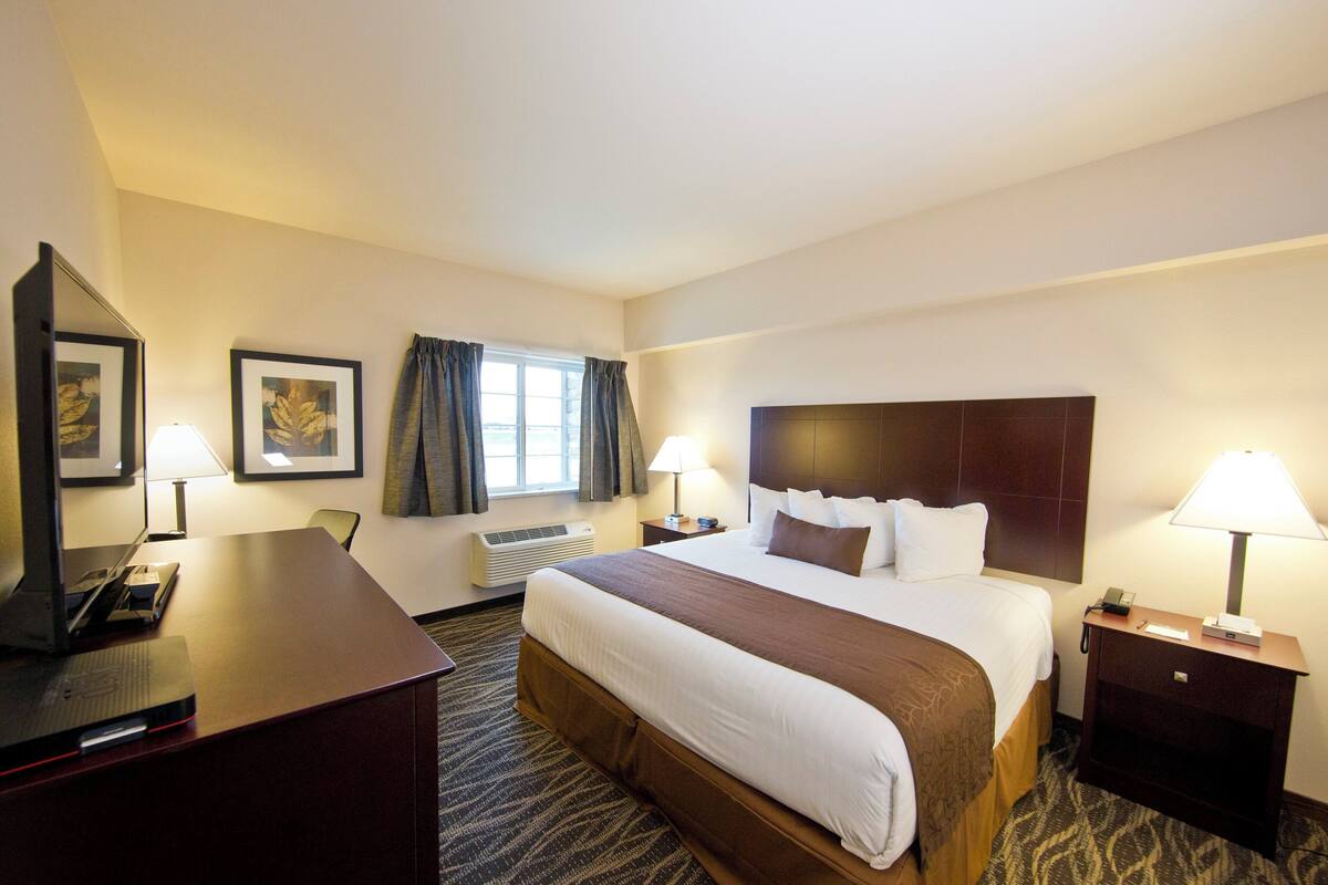 A hotel room with a large bed, two bedside tables with lamps, a desk with a computer, a window with curtains, and framed artwork on the wall.
