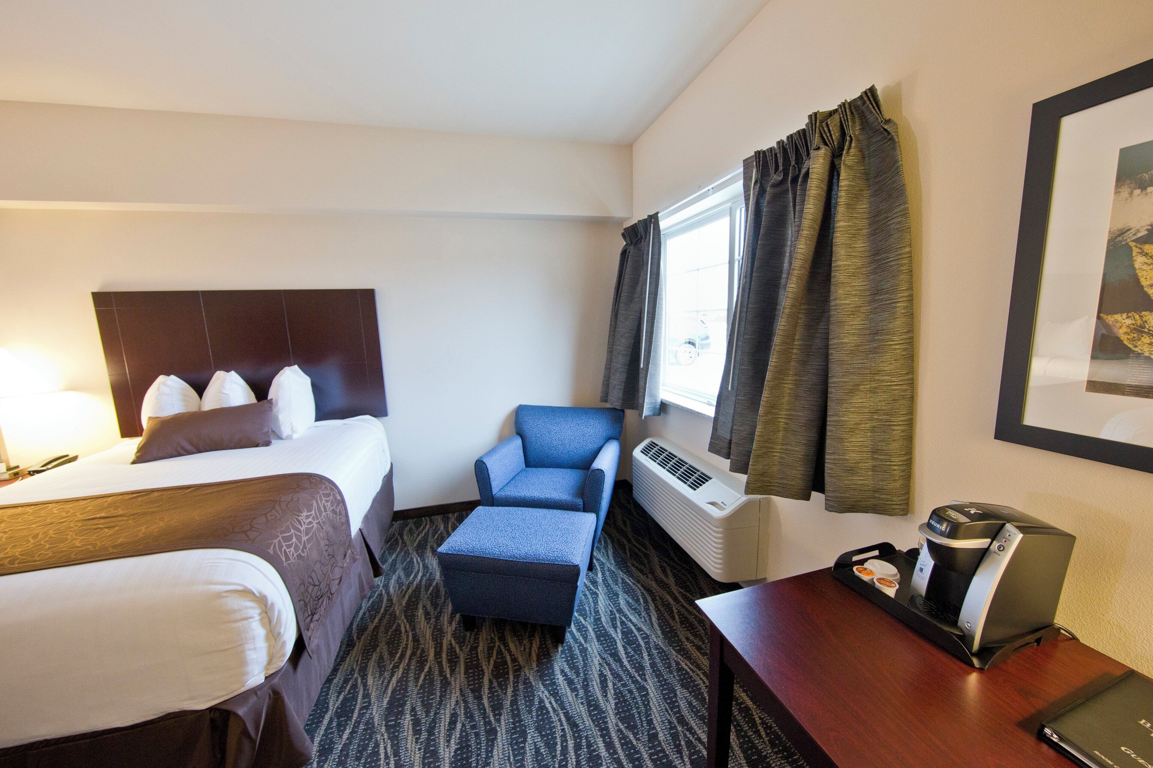 A hotel room with a bed, a chair, a desk with a coffee maker, and a window with curtains.