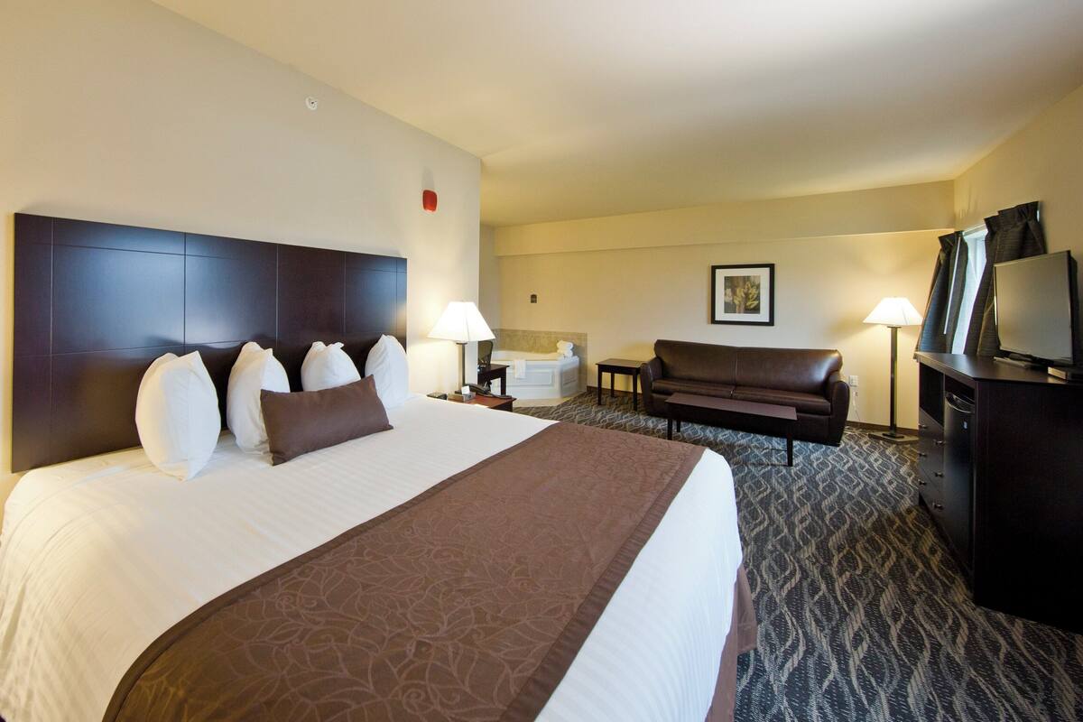 A hotel room with a large bed, a sofa, a TV, a desk, and a bathroom in the background.