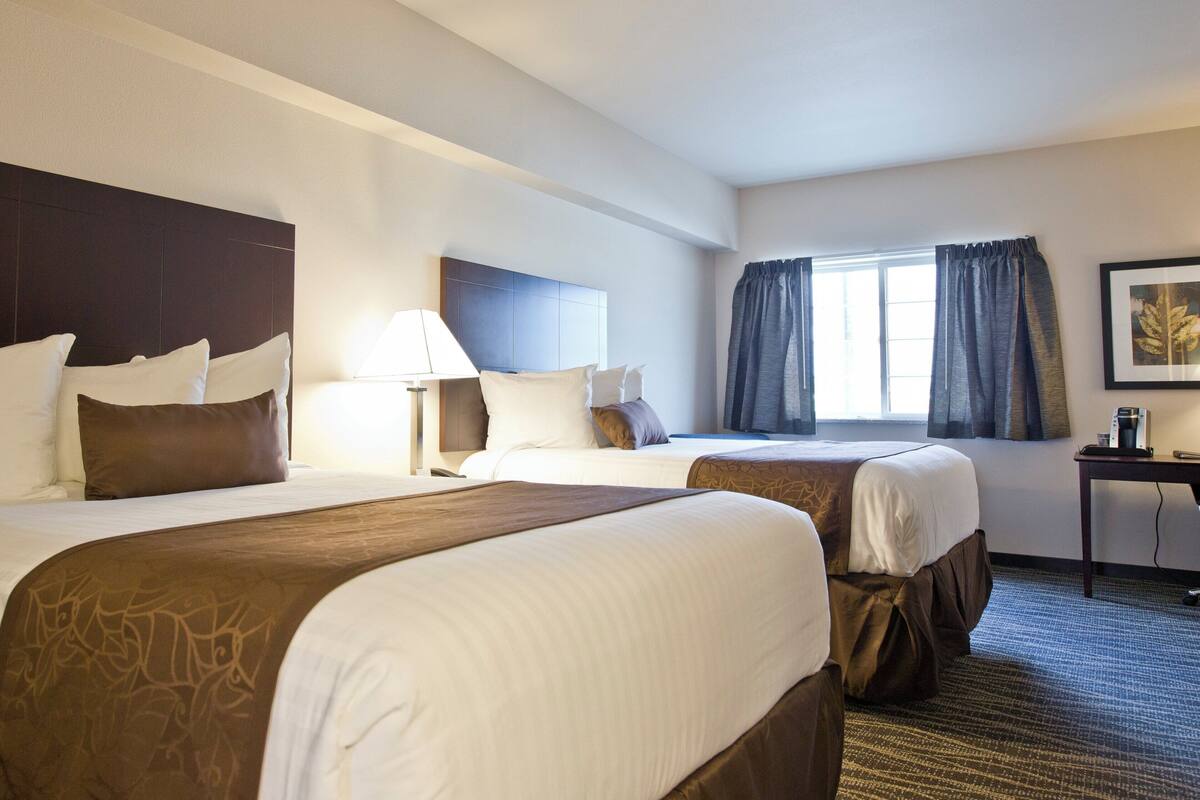 A hotel room with two beds, a desk, a lamp, and a window with curtains.