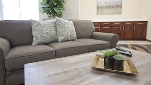 Uma sala de estar com um sofá cinza, almofadas estampadas, uma mesa de centro com um tabuleiro contendo plantas em vasos, um controle remoto e um quadro na parede.