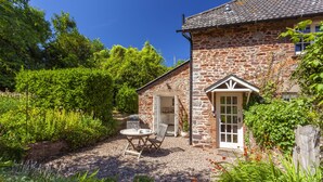A stone building with a gravel patio, a small garden, and a door leading to a covered area. - Horner Cottage, Luccombe (Nr Porlock)