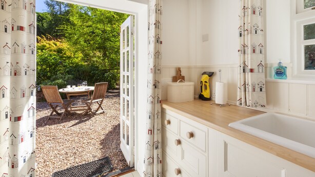 A kitchen with a wooden countertop, white cabinets, and a view of a garden with chairs and a table. - Horner Cottage, Luccombe (Nr Porlock)
