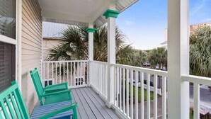 A covered porch with white railings and green accents, featuring two green chairs and a view of a residential area with palm trees and a white picket fence. - Eden's Estate - Pet Friendly, Heated Community Pool, Bikes Included! (Miramar Beach)