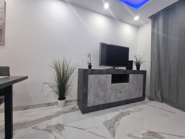 Un salon moderne avec un sol en marbre, un meuble de rangement gris intégrant un téléviseur à écran plat, et deux plantes en pot.