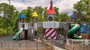 A playground with multiple slides and climbing structures, surrounded by a wooden fence and trees.