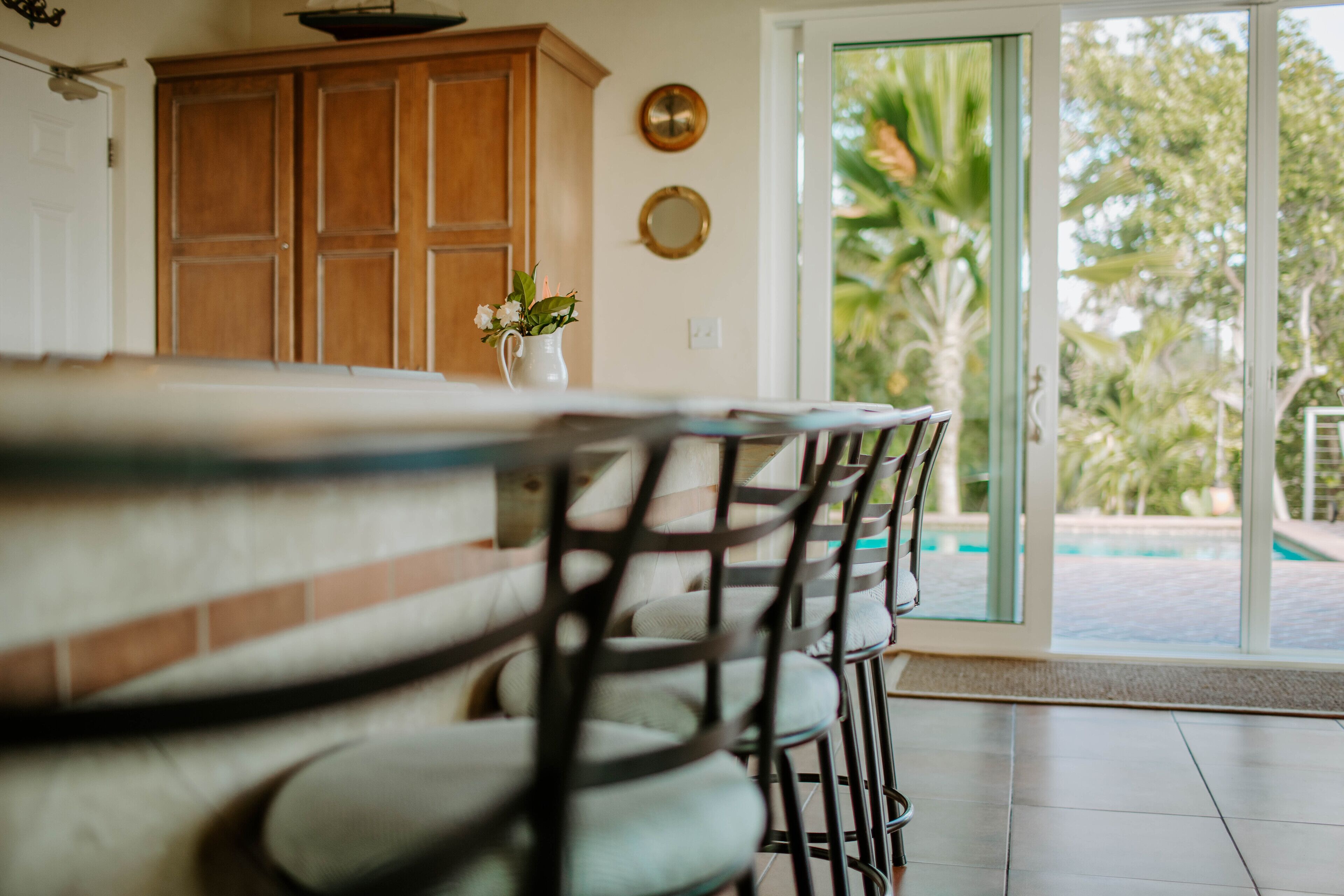 A kitchen with a wooden cabinet, a white vase with flowers, and a view of a swimming pool through glass doors.