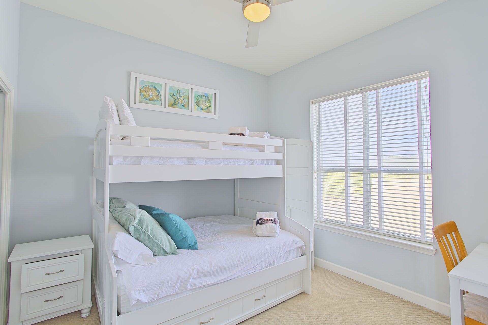 Una cama litera con escritorio y silla, una ventana con persianas, y cuadros en la pared.