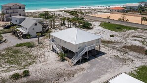 Una propiedad frente a la playa con un edificio central que presenta una escalera que lleva a una terraza, rodeada de terreno arenoso y vegetación escasa.