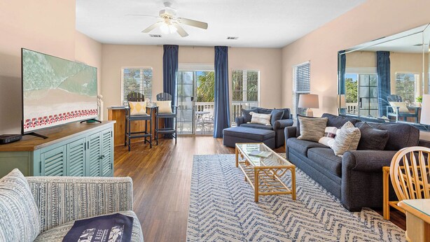 A spacious living room with a large flat-screen TV, a ceiling fan, and a view of the outdoors through large windows. - Legends 310 - Santa Rosa Beach, Community Pool, Hot Tub, Tennis Courts! (Santa Rosa Beach)