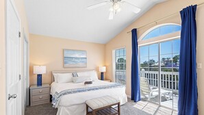 A bedroom with a bed, bedside tables, a bench, a ceiling fan, a window with curtains, and a view of the outside. - Legends 310 - Santa Rosa Beach, Community Pool, Hot Tub, Tennis Courts! (Santa Rosa Beach)