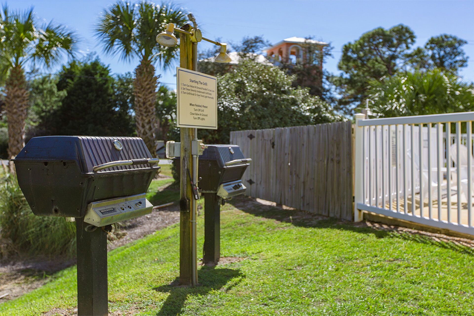 Two barbecue grills with a sign indicating grilling instructions, a wooden fence, and a building in the background.