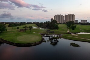 Campo de golf con una masa de agua, un puente y altos edificios residenciales al fondo.