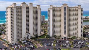 Two high-rise buildings with multiple balconies, a parking lot, and a view of the ocean.