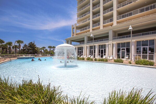 A swimming pool with a central fountain, surrounded by lounge chairs and a multi-story building with balconies.