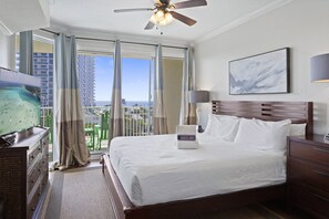 Quarto de hotel com cama, televisão, ventoinha no teto, mesa de cabeceira, abajur e vista para o oceano e edifícios.