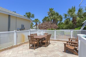 Une terrasse avec une table et des chaises en bois, entourée d’une clôture blanche, avec des palmiers et un bâtiment en arrière-plan.