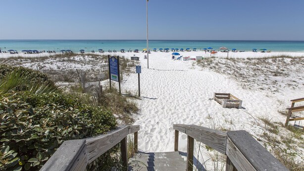 Une plage avec une promenade en bois menant à l’eau, des parasols de plage et un mât de drapeau.
