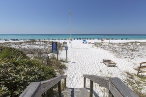 En strand med en träpromenad som leder ner till vattnet, strandparasoller och en flaggstång.