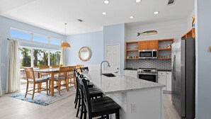 A modern kitchen with a central island, wooden dining chairs, and a view of the outdoors. - Hideout – Updated! Pet Friendly & Walk to Beach! (Destin)