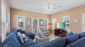 A living room with a blue sofa, a grey armchair, a TV, and a ceiling fan. - All 4 Fun - Gulfside Cottages, Community Pool, Steps to the Beach! (Miramar Beach)