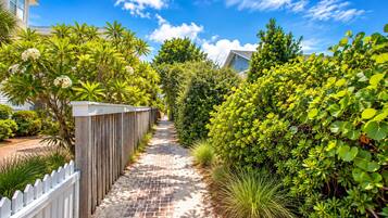 A pathway flanked by lush greenery and a wooden fence, leading to a building with a white facade.