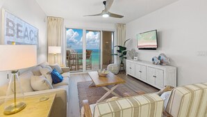 A spacious living room with a balcony, a ceiling fan, a television, and a view of the ocean. - Ariel Dunes I 1902 - Gulf Views! Community Pool! Onsite Dining! Exercise Room! (Miramar Beach)