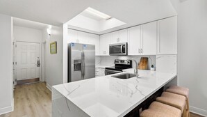 A modern kitchen with white cabinetry, a large island with bar stools, stainless steel appliances, and a skylight. - Ariel Dunes I 1902 - Gulf Views! Community Pool! Onsite Dining! Exercise Room! (Miramar Beach)