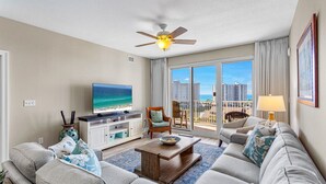A modern living room with a flat-screen TV, comfortable seating, a ceiling fan, and a view of the ocean and buildings. - Ariel Dunes II 707 - Gulf Views! Heated Community Pool! Fitness Center! Book Now (Miramar Beach)