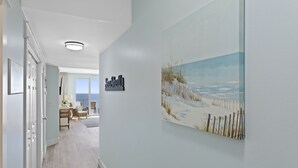 Un couloir orné d’un tableau représentant une scène de plage, une pancarte indiquant « Coquillages », et une vue sur un balcon avec une chaise et une table.