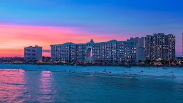Un hotel grande con varios pisos y un patio central, situado cerca de una playa con gente en la arena y agua.
