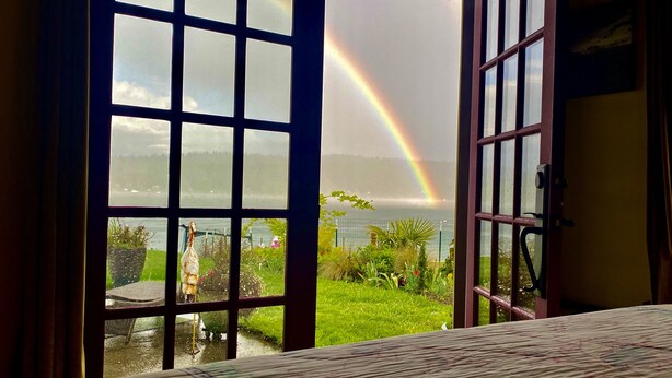 A room with a bed, a view of a garden, and a rainbow visible through a window. - SHABBY CHIC WATERFRONT RETREAT serene low bank waterfront home w/ gas fire pit. (Vashon)