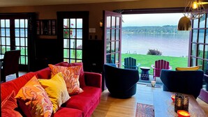 A cozy living room with a red sofa, a black chair, and a table with candles. Large windows offer a view of a lake and greenery. - SHABBY CHIC WATERFRONT RETREAT serene low bank waterfront home w/ gas fire pit. (Vashon)