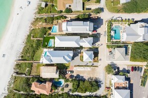 Vista aérea de un complejo turístico costero con varios edificios, incluyendo un gran edificio principal con piscina, casas de huéspedes más pequeñas y una zona de playa con sombrillas.