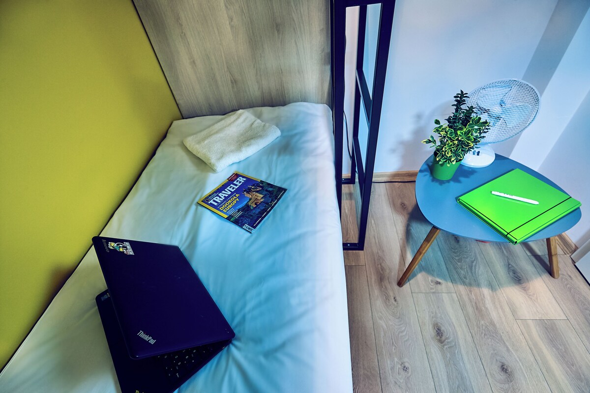 Ett litet, välordnat rum med en säng, en bärbar dator, en tidning, ett grönt anteckningsblock och ett litet bord med en fläkt och en växt.