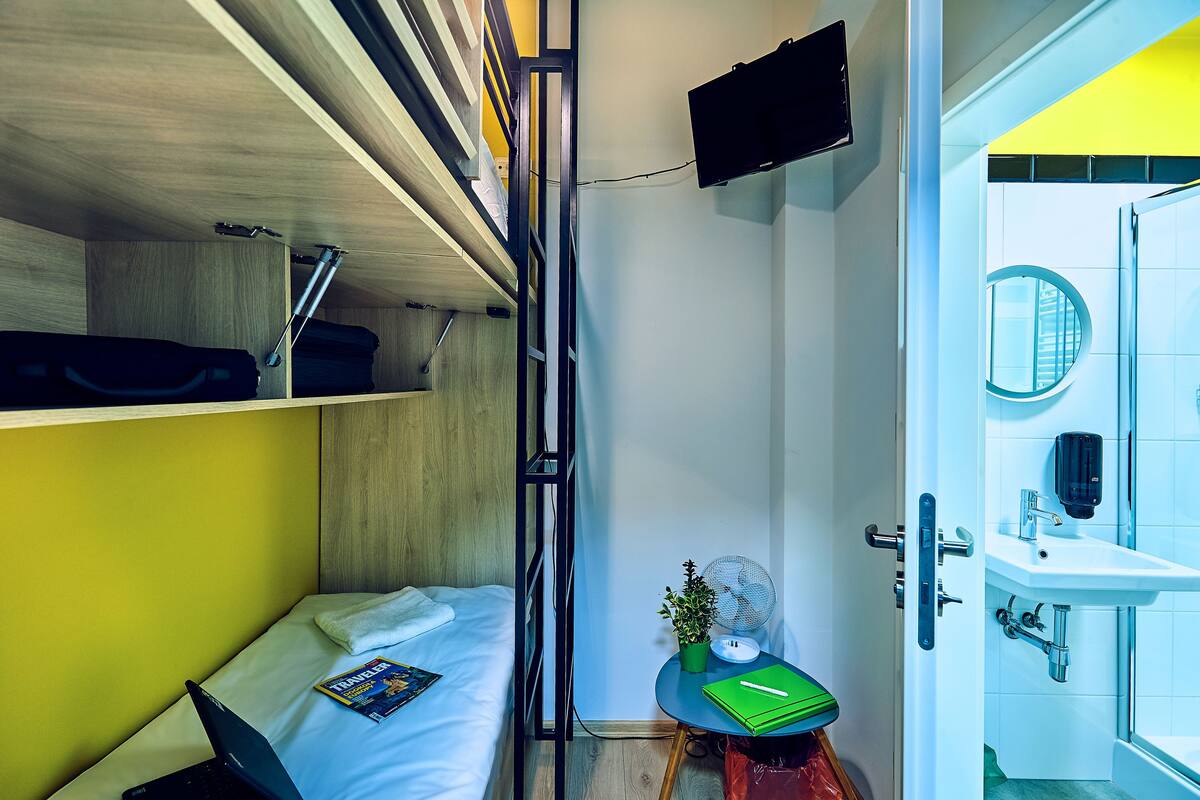 A compact hotel room with a bunk bed, a small table with a plant, a laptop, and a magazine on the bed.