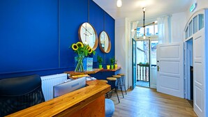 A reception area with a wooden desk, black office chair, and a blue wall with two round mirrors. There are potted plants on a shelf and a sign with text. - Draggo House- Hostel (Kraków)