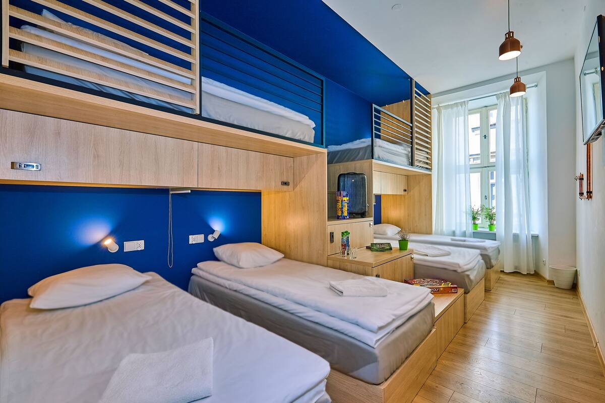 A modern hotel room with three beds, a wooden wardrobe, a small table with a lamp, and a window with curtains.