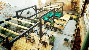 An outdoor patio area with wooden tables and chairs, a green sofa, and hanging plants. - Draggo House- Hostel (Kraków)