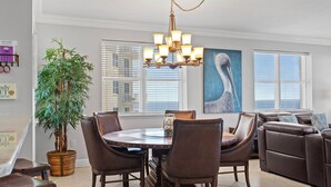 A dining area with a round table, chairs, a chandelier, a large potted plant, and a painting on the wall.