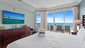 A hotel room with a large bed, a desk with a computer, a chair, a TV on the wall, and a view of the ocean.