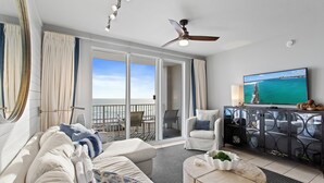 A modern living room with a large TV, a ceiling fan, and a view of the ocean through the sliding glass doors. - Majestic Sun 208B - Upscale Unit, Gulf Views, Community Pool + Hot Tub! (Miramar Beach)