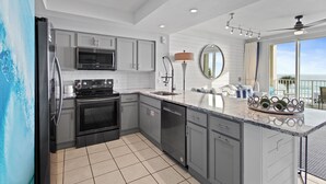 A modern kitchen with grey cabinets, stainless steel appliances, a large island with a sink, and a view of the ocean through a large window. - Majestic Sun 208B - Upscale Unit, Gulf Views, Community Pool + Hot Tub! (Miramar Beach)