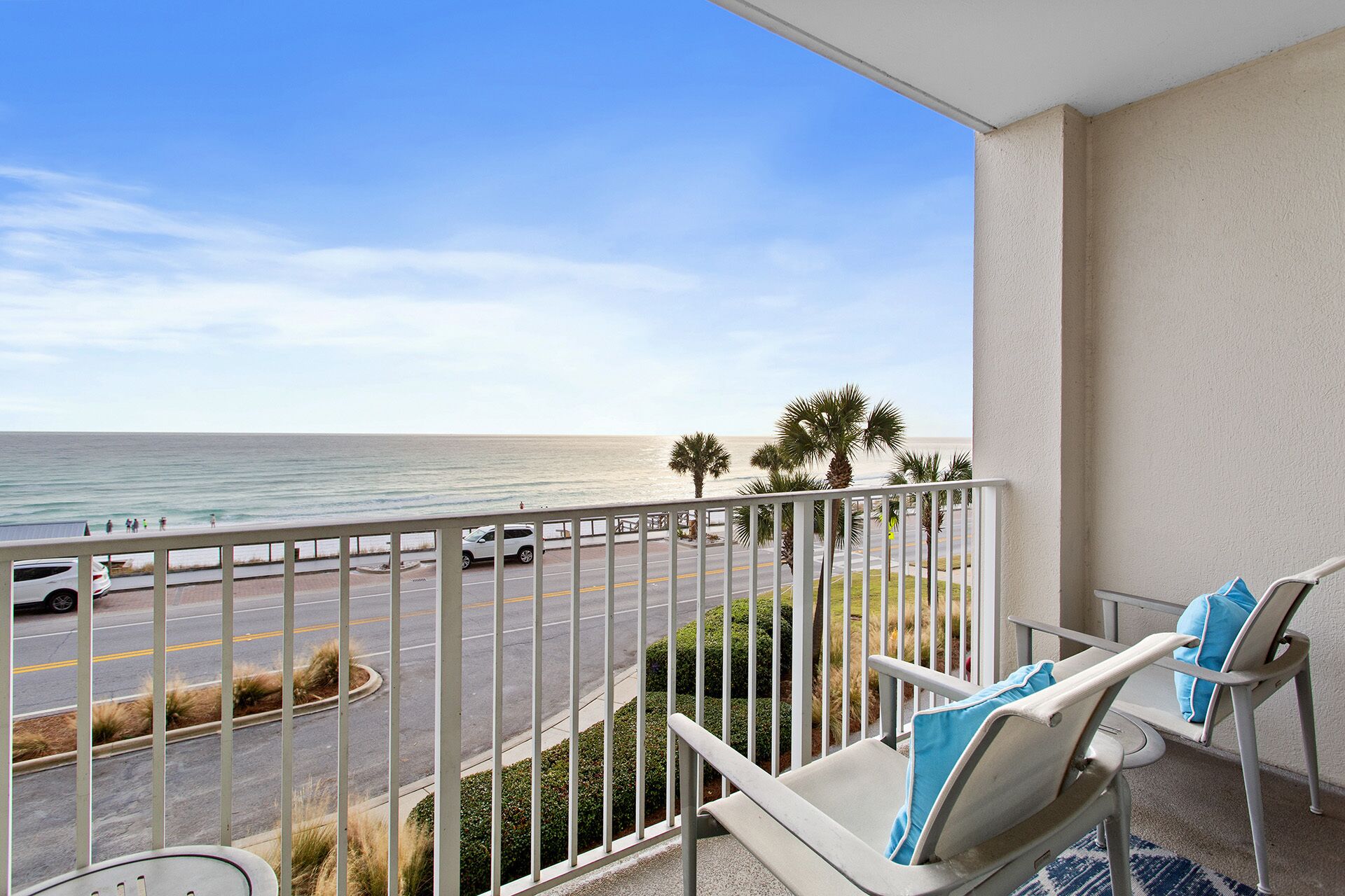 A balcony with a view of the ocean, a parking lot, and a street with palm trees.