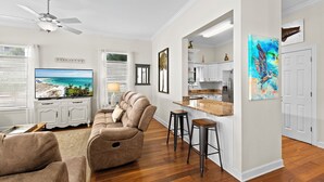 A living room with a large flat-screen TV, a sofa, a ceiling fan, and a kitchen area with white cabinets and a painting. - Sandy By The Sea - Blue Mountain Beach! Gulf View! Private Pool! Pet Friendly! (Santa Rosa Beach)
