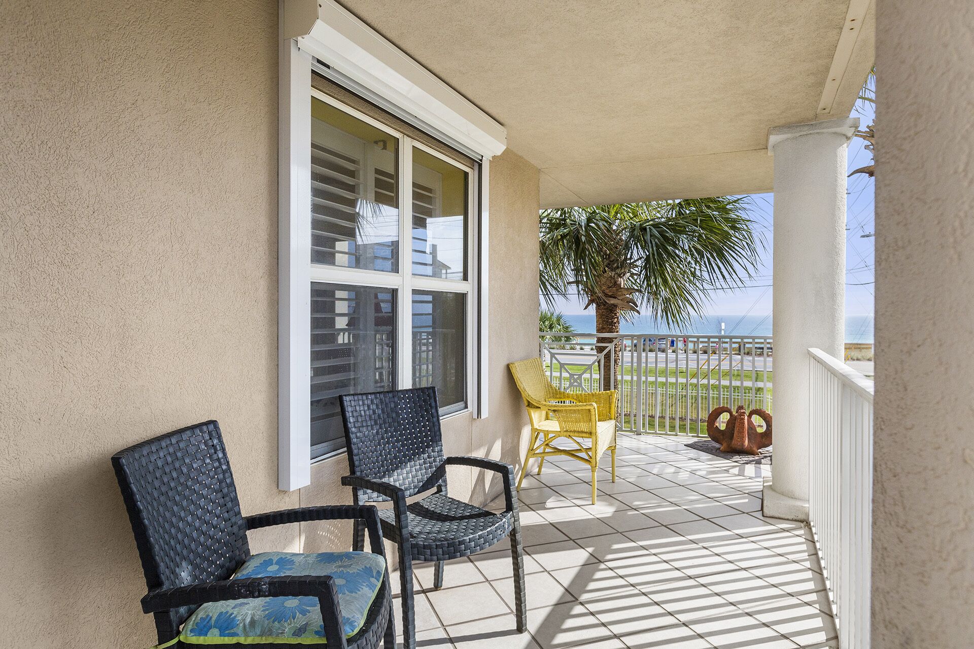 A balcony with two wicker chairs, one with a blue cushion and another with a yellow cushion, and a large window with blinds.