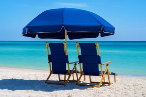 Deux chaises de plage sous un parasol bleu, sur une plage de sable fin bordée d’une eau turquoise et cristalline.